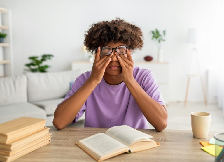 Black teen guy rubbing tired eyes, sitting at desk with open textbook, exhausted from home schooling